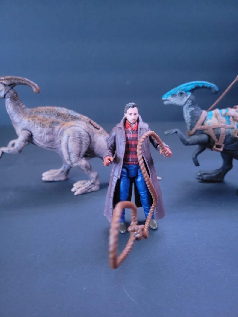 Pu&ograve; includere: Una action figure di un uomo che indossa una giacca marrone, una camicia a righe rosse e bianche, jeans blu e tiene una corda marrone. La figura &egrave; in piedi davanti a due action figure di dinosauri, una marrone e una blu.
