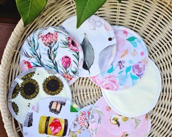 May include: A selection of round, reusable nursing pads featuring diverse floral and patterned designs, such as sunflowers, protea flowers, and watercolour illustrations. The pads are displayed in a woven basket, with green leaves visible.