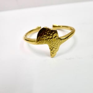 Puede incluir: Un anillo dorado con un diseño texturizado en forma de África. El anillo es abierto en la parte trasera, lo que permite ajustar la talla. El anillo está sobre un fondo blanco.