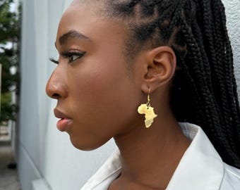 May include: Gold-colored earrings in the shape of the African continent. The earrings are hanging from a person's ear. The person is wearing a white collared shirt and has their hair in braids. The background is a white wall.