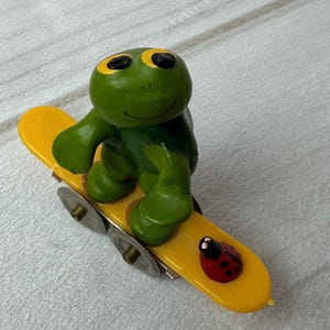 May include: A whimsical green frog figurine with black eyes and yellow accents, riding a yellow skateboard. A red ladybug with black spots is on the skateboard. The skateboard has four small wheels.