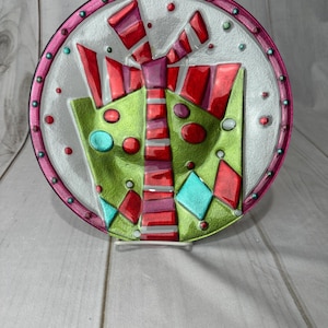 May include: A decorative glass plate featuring a colorful gift box design. The plate has a clear center with a green gift box, red bow, and accents of teal and red. The rim is pink with small, colorful embellishments.
