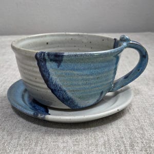 May include: A ceramic cup and saucer set featuring a speckled off-white base with horizontal ridges. The exterior of the cup and the edge of the saucer are decorated with a deep blue glaze that drips down the side, complemented by a curved handle.
