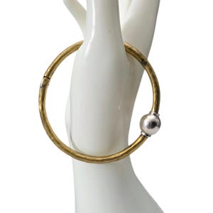 May include: A gold-colored metal bangle bracelet with a silver-colored ball clasp. The bracelet is round and appears to be made of hammered metal. The bracelet is displayed on a white hand.