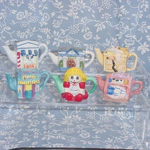 May include: A collection of six decorative ceramic teapots, each uniquely designed with vibrant colors and intricate details. The teapots are arranged on a clear, tiered display stand against a light blue floral background.