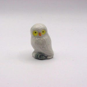 May include: A small white ceramic owl figurine with yellow eyes and an orange beak. The owl is perched on a green base.