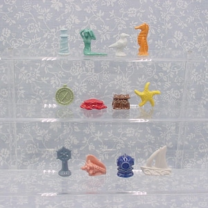 May include: A collection of miniature ocean-themed figurines, including a lighthouse, mermaid, bird, seahorse, compass, crab, treasure chest, starfish, ship's wheel, seashell, diving helmet, and sailboat. The figurines are in various colors and arranged on clear shelves.