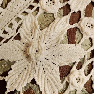 May include: Close-up of a white, handmade crochet doily featuring a floral design. The doily has a detailed pattern of flowers and leaves, with openwork sections. The background is a mix of brown and green fabric.