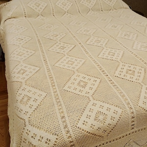 May include: Cream-colored, intricately crocheted bedspread with a geometric diamond pattern. The handmade blanket features detailed openwork and a scalloped edge, adding a touch of vintage charm to any bedroom decor.