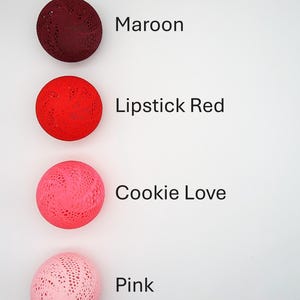 May include: Four decorative balls in different shades of red and pink. The balls are made of a lacy material and are arranged vertically from darkest to lightest. The colors are labeled as Maroon, Lipstick Red, Cookie Love, and Pink.