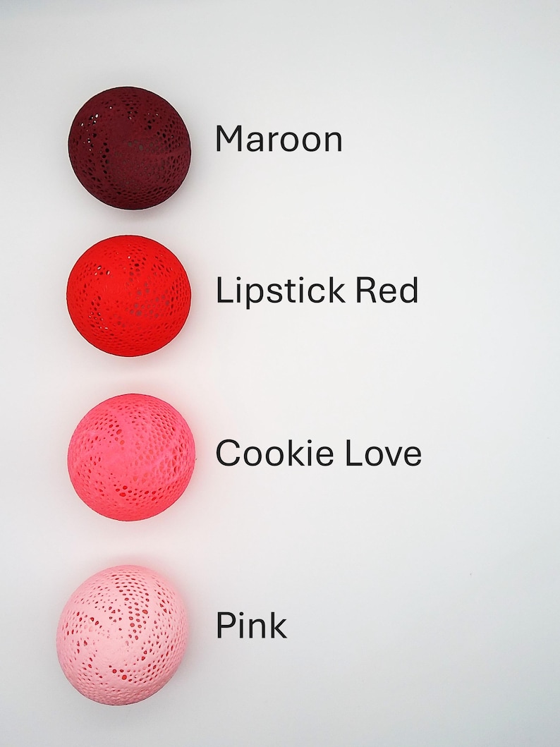 May include: Four decorative balls in different shades of red and pink. The balls are made of a lacy material and are arranged vertically from darkest to lightest. The colors are labeled as Maroon, Lipstick Red, Cookie Love, and Pink.
