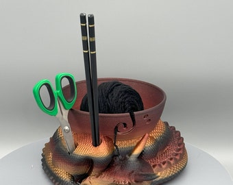 May include: A ceramic yarn bowl with a dragon design in shades of red, orange, and black. The bowl has a hole for yarn to feed through and is holding a ball of black yarn. Two black chopsticks are standing upright in the bowl, and a pair of green scissors are resting on the dragon's head.