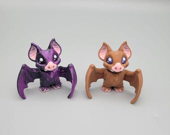 Könnte beinhalten: Zwei kleine, 3D-gedruckte Fledermausfiguren. Eine ist lila mit schwarzen Flügeln und die andere ist braun mit braunen Flügeln. Beide haben rosa Ohren und Nasen.