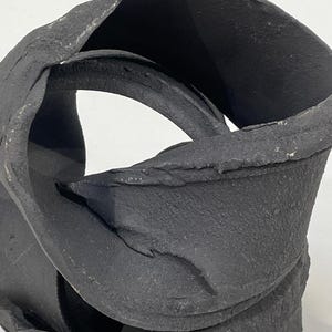 May include: A black, abstract ceramic sculpture with a textured surface. The sculpture has a looped, intertwined design, with a central opening. The piece is set against a white background, highlighting its form and texture.