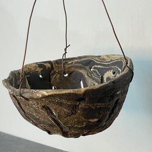 May include: A hanging planter with a marbled design in shades of brown and cream. The ceramic pot has cut-out details and is suspended by wire hangers. Pieces of the same material are scattered below.