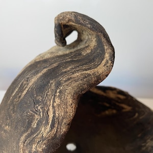 May include: Close-up of a handcrafted, organic-shaped ceramic bowl. The bowl features a dark brown and tan marbled pattern, with a curled rim and a small hole near the base. The texture appears rough and natural.