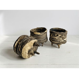 May include: Three handmade ceramic planters with a unique, textured design. The planters feature a blend of brown and cream tones, with a marbled effect. Each planter has a cylindrical shape and rests on three small legs.