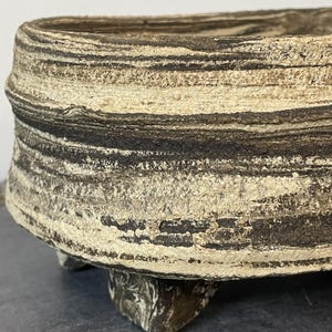 Puede incluir: Una jardinera ovalada hecha a mano con un diseño texturizado en capas. La jardinera presenta bandas alternas de marrón oscuro y blanquecino, creando una estética natural. Descansa sobre tres pequeños pies redondeados.