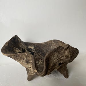 Könnte beinhalten: Eine dunkelbraune, organisch geformte Schale aus Naturholz. Die Schale hat eine raue, strukturierte Oberfläche mit sichtbarer Holzmaserung, tief geschwungene Ränder und drei kurze, stabile Füße, die den Boden des Gefäßes stützen.