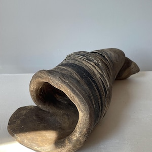 May include: A sculptural ceramic object with a dark, earthy, and marbled appearance. The piece features a hollow, rolled, and elongated form with visible layers of black, gray, and brown tones, creating a textured, organic, and swirling surface.