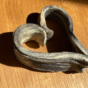 Peut inclure: Un objet décoratif en forme de cœur avec un aspect texturé et vieilli. La sculpture est principalement grise avec des stries beige clair, créant une esthétique naturelle semblable à la pierre. Posé sur une surface en bois.