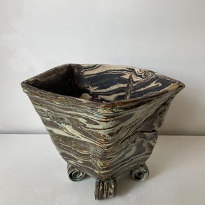 Peut inclure: Un pot de fleurs carré en céramique présentant un motif marbré de tourbillons brun foncé, noirs et blanc cassé. Le récipient possède un bord large et évasé, soutenu par quatre petits pieds enroulés à la base, avec un aspect texturé.