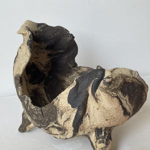 Może przedstawiać: Ręcznie wykonana ceramiczna misa o nieregularnym, organicznym kształcie i teksturowanej powierzchni. Przedmiot posiada marmurkowy wzór w kolorach matowej czerni i złamanej bieli, wsparty na trzech krótkich, solidnych nóżkach unoszących podstawę.