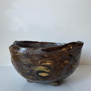 Könnte beinhalten: Eine handgeformte Keramikschale in Dunkelbraun mit einer glänzenden, unebenen Glasur und einer strukturierten, rauen Oberfläche. Die Schale hat einen breiten, unregelmäßigen Rand und steht auf kleinen, integrierten Füßen in tiefen Brauntönen.