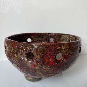 May include: A round, footed ceramic bowl with a textured, dark brown and metallic gold finish featuring vibrant red glaze drips. The bowl has a series of evenly spaced circular cutouts around the upper rim, creating a decorative, openwork design.