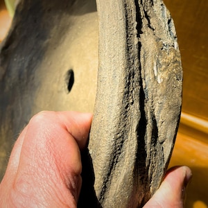 Könnte beinhalten: Eine kreisförmige, braune und texturierte Tonschale mit einem dicken, unebenen Rand und einem kleinen Abflussloch in der Mitte. Die Oberfläche der Schale weist ein raues, verwittertes Aussehen mit sichtbaren Rillen und einem matten Finish auf.