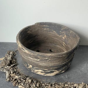 Puede incluir: Una maceta redonda de cerámica hecha a mano con un diseño veteado marrón oscuro y crema. La maceta tiene una abertura ancha y dos orificios de drenaje. Las virutas de madera decorativas rodean la base de la maceta.