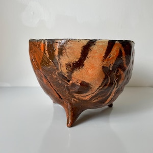 May include: A hand-built ceramic bowl with a tripod base, featuring a marbled glaze in shades of deep brown, burnt orange, and cream. The exterior surface has a textured, uneven finish with visible indentations and a glossy, reflective sheen.