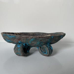 May include: A hand-built ceramic bowl with an irregular, textured surface in dark brown and vibrant blue. The bowl features a wide, shallow basin and is supported by three distinct, coiled, scroll-shaped feet that match the bowl's color palette.