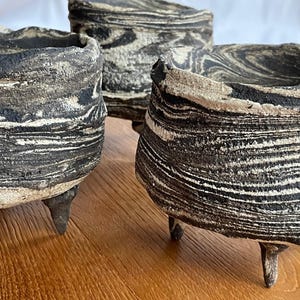 Peut inclure: Trois pots en céramique faits à la main avec un design marbré unique en noir et blanc. Chaque pot a une forme arrondie et est soutenu par trois petits pieds pointus. Les pots sont exposés sur une surface en bois.