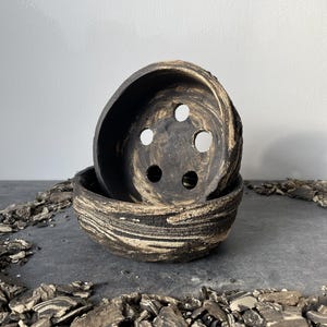 Puede incluir: Dos cuencos de cerámica marrón oscuro hechos a mano con un diseño veteado único. El cuenco superior tiene orificios de drenaje. Los cuencos están apilados uno encima del otro. Los cuencos están rodeados de trozos de corteza.