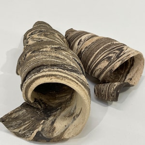 May include: Two decorative ceramic sculptures with a spiral seashell design. The sculptures are brown and beige with a marbled pattern. The open ends of the sculptures are visible, revealing the interior. The pieces are on a white surface.