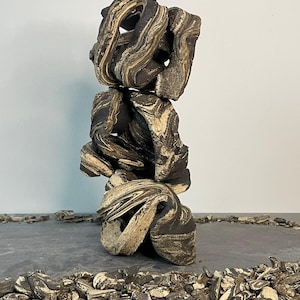 May include: A stacked sculpture composed of dark brown and tan swirled stone. The sculpture has a unique, organic shape with open spaces. Small stone fragments are scattered around the base, adding texture and detail to the display.