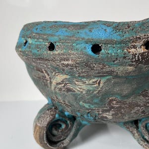 Op de afbeelding: Een donkerbruine keramische kom met een ruw, getextureerd oppervlak en levendige blauwe glazuuraccenten. De kom heeft een reeks kleine, gelijkmatig verdeelde gaatjes langs de bovenrand en staat op drie opvallende, gekrulde, spiraalvormige voetjes.