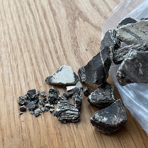 May include: A collection of small, irregularly shaped rocks with a mix of black and white striped patterns. Some rocks are solid black or white. The rocks are scattered on a wooden surface, with some spilling from a clear plastic bag.