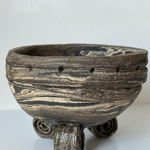 May include: A hand-built ceramic bowl featuring a marbled black and beige clay pattern. The bowl has a thick, textured rim with a series of small, evenly spaced indentations and is supported by three distinct, coiled, scroll-shaped feet.