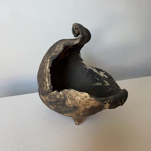 May include: A handcrafted, abstract ceramic bowl with a unique, organic shape. The bowl features a dark, textured exterior with a lighter, golden-brown interior and rim. It rests on three small feet.