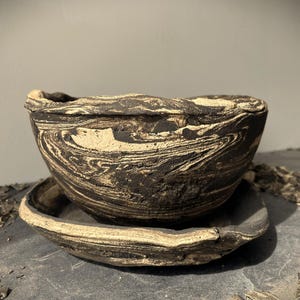 May include: A handcrafted ceramic bowl and saucer set. The bowl is dark brown with a marbled pattern of cream and black. The saucer has a similar design. The set is suitable for serving food or as a decorative piece.