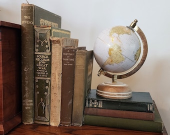 Puede incluir: Una colección de libros antiguos con títulos como "The Studio", "Source Records of the Great War" y "A History of Painting" están apilados en un estante de madera junto a un pequeño globo terráqueo.