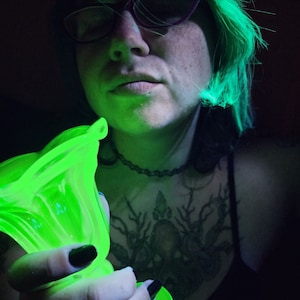 May include: A person with green hair holds a green glass vase with a fluted design. The vase is illuminated with a green light, creating a glowing effect.