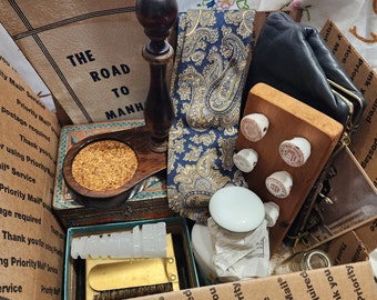 May include: A brown cardboard box filled with various items, including a wooden stamp, a paisley patterned tie, a small wooden coaster, a white ceramic jar, and a brown leather wallet. The box is labeled with the text "Priority Mail" and "The Road to Manhattan".