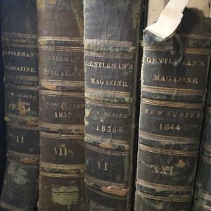 May include: Four antique books titled Gentleman's Magazine New Series with years 1834, 1837, 1836, and 1844 printed on the spines. The dark, weathered leather spines feature gold lettering, raised bands, and visible signs of age and wear.