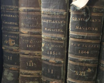 Op de afbeelding: Vier antieke boeken getiteld Gentleman's Magazine New Series met de jaartallen 1834, 1837, 1836 en 1844 op de ruggen. De donkere, verweerde lederen ruggen zijn voorzien van gouden letters, ribben en vertonen duidelijke sporen van ouderdom.