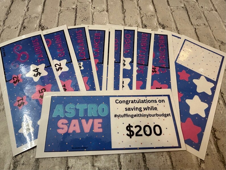 May include: A set of 10 colorful cards with astrological signs and dollar amounts. The cards are arranged in a fan shape. The bottom card says "ASTRO SAVE" and "Congratulations on saving while #stuffingwithinyourbudget $200".