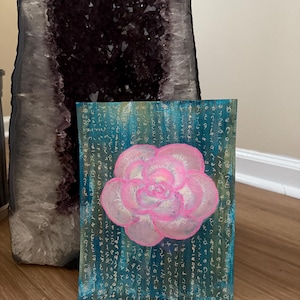 May include: A pink flower painted on a teal canvas with white and gold script. The canvas is leaning against a large amethyst geode.