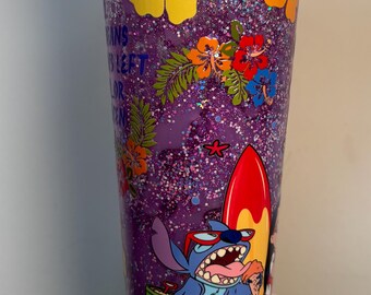 May include: Purple glitter tumbler with a cartoon character design. The tumbler features a blue character wearing sunglasses, a red and yellow surfboard, and colorful floral accents. The tumbler has a clear lid.